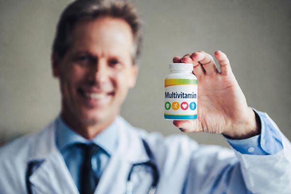 Fehér köpenyes orvos mosolyogva tart a kezében egy doboz multivitamint.