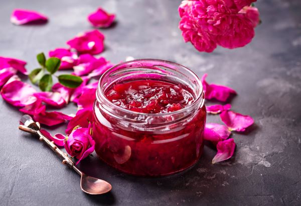 Egy kis üveg rózsalekvár mellett rózsaszirmok és egy kiskanál.
