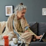Idős nő a kanapén ülve nézegeti a tabletjét, vállára terítve plédek, előtte az asztalon gyógyszerek.