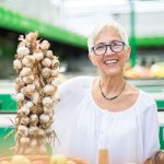 Középkorú szemüveges nő a piacon nagy füzér fokhagymát tart a kezében mosolyogva,