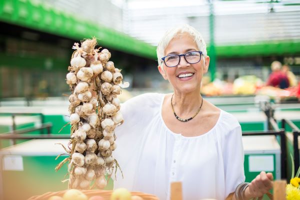 Középkorú szemüveges nő a piacon nagy füzér fokhagymát tart a kezében mosolyogva,