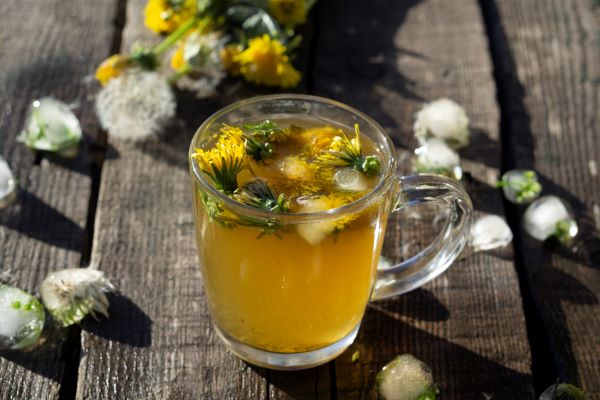Rusztikus faasztalon füles üvegbögrében pitypangtea, mellette egy csokor pitypang.