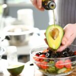 Valaki olívaolajat csurgat egy fél avokádóra egy üvegtál fölött, amelyben olívabogyó, paradicsom, uborka van felszeletelve.