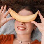 Nő egy pléden fekszik és mosolyogva a szemei elé tart egy banánt.