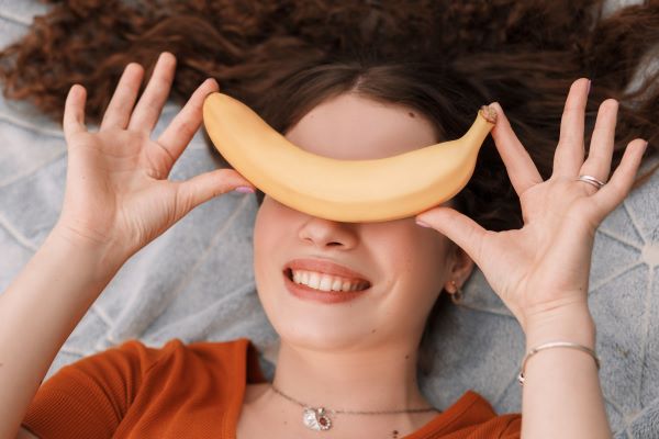 Nő egy pléden fekszik és mosolyogva a szemei elé tart egy banánt.