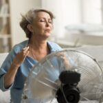 Középkorú nő ül a kanapén a ventilátor előtt.
