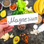 Egy kéz tart egy papírt "Magnesium" felirattal, a háttérben magnéziumban gazdag táplálékok.