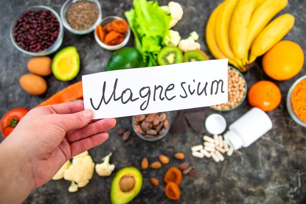 Egy kéz tart egy papírt "Magnesium" felirattal, a háttérben magnéziumban gazdag táplálékok.