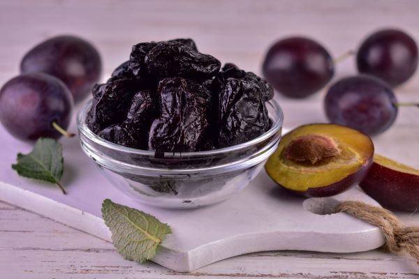 Fehér vágódeszkán kis üvegtálban aszalt szilva, mellette néhány szem friss szilva.