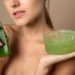 Fiatal félmeztelen nő egyik kezében egy Aloe vera levelet, másikban egy tégely Aloe vera gélt tart.