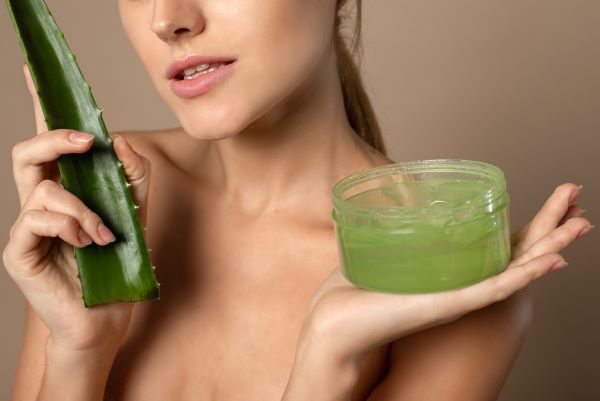 Fiatal félmeztelen nő egyik kezében egy Aloe vera levelet, másikban egy tégely Aloe vera gélt tart.