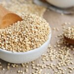 Fehér porcelántálban quinoa fakanállal, mellette az asztalon fakanál quinoával.