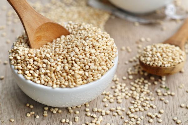 Fehér porcelántálban quinoa fakanállal, mellette az asztalon fakanál quinoával.