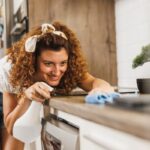Nő takarítja a konyhapultot, egyik kezében törlőkendő, másikban szórófejes flakon.