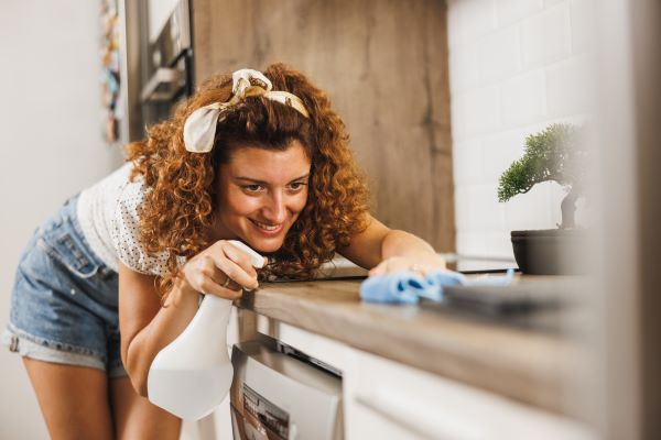 Nő takarítja a konyhapultot, egyik kezében törlőkendő, másikban szórófejes flakon.