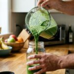 Férfi a konyhában a turmixgép kancsójából zöld turmixot tölt egy befőttesüvegbe.