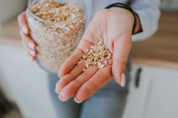 Nő egyik tenyerében kevés zabpelyhet tart oda a kamerának, másik kezében egy üveg zabpehely.