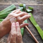 Idősebb nő Aloe vera gélt tesz az egyik kézfejére egy rusztikus faasztal fölött, amelyen Aloe vera levelek hevernek.