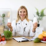 Fiatal doktornő az asztalnál ülve mosolyogva tart mindkét kezében egy-egy étrend-kiegészítős dobozt, előtte az asztalon gyümölcsök és zöldségek.