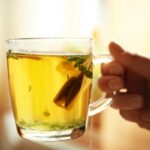 Női kézben egy pohár tea teafilterrel és gyógynövénnyel.