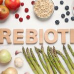 Milyen előnyei vannak a prebiotikumoknak? "Prebiotic" szó kirakva, körülötte gyümölcsök és zöldségek: alma, áfonya, málna, zabpehely, paradicsom, hagyma, fokhagyma, spárga.