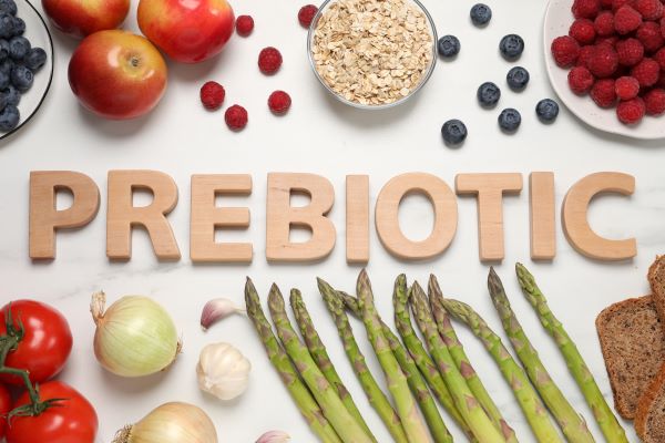 "Prebiotic" szó kirakva, körülötte gyümölcsök és zöldségek: alma, áfonya, málna, zabpehely, paradicsom, hagyma, fokhagyma, spárga.