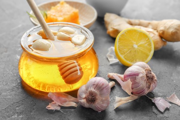 Egy üvegmáz fokhagymagerezdekkel, mellette két fej fokhagyma, egy gyömbér és egy fél citrom.
